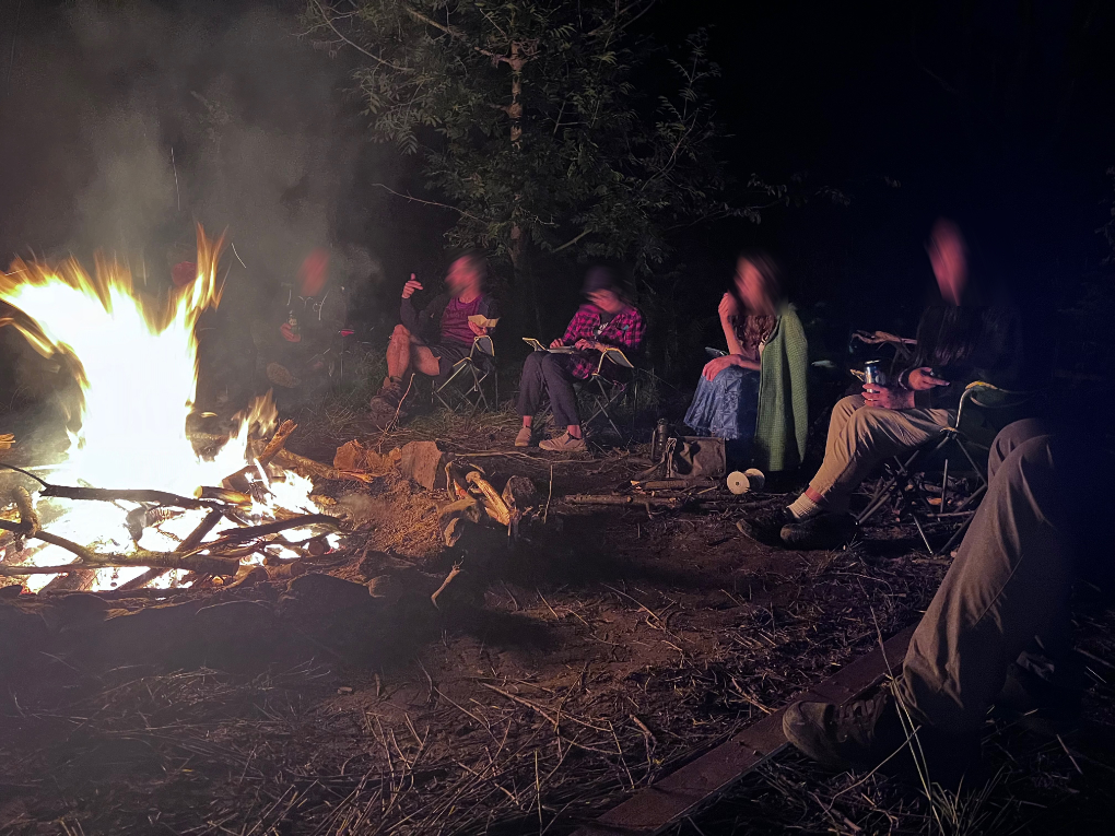 On the left a camp fire burning brightly, on the right attendees sit in chairs around the fire